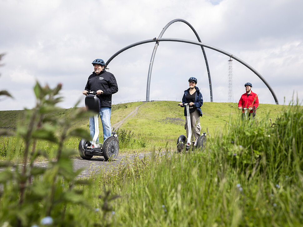 Zwei Segwayfahrer folgen einem Guide auf einer Tour über die Halde Hoheward in Herten. Im Hintergrund ist das Horizontobservatorium zu sehen.