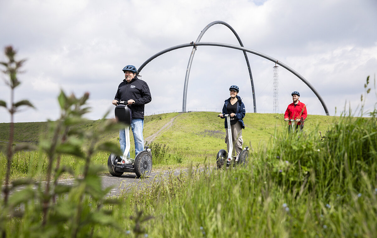 Zwei Segwayfahrer folgen einem Guide auf einer Tour über die Halde Hoheward in Herten. Im Hintergrund ist das Horizontobservatorium zu sehen.