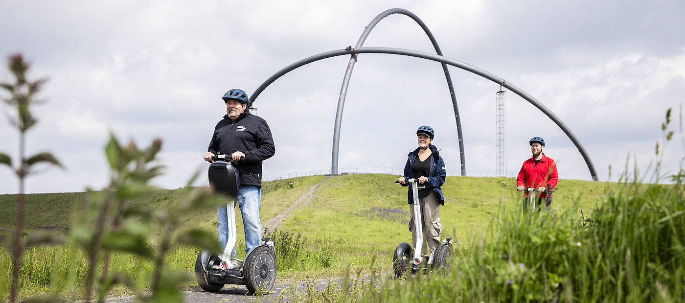 Zwei Segwayfahrer folgen einem Guide auf einer Tour über die Halde Hoheward in Herten. Im Hintergrund ist das Horizontobservatorium zu sehen.