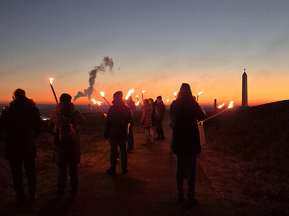 Eine Gruppe Menschen mit Fackeln läuft im Sonnenuntergang Richtung Sonnenuhr der Halde Hoheward.