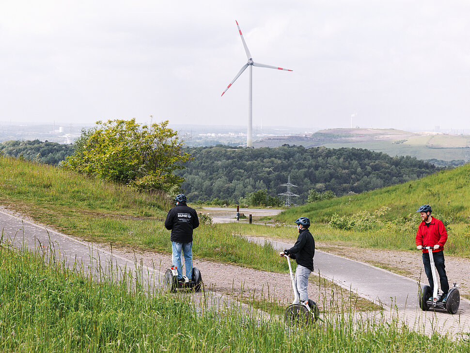 Zwei Segwayfahrer folgen einem Guide auf einer Tour über die Halde Hoheward in Herten. Im Hintergrund ist die Halde Hoppenbruch mit dem Windrad zu sehen.