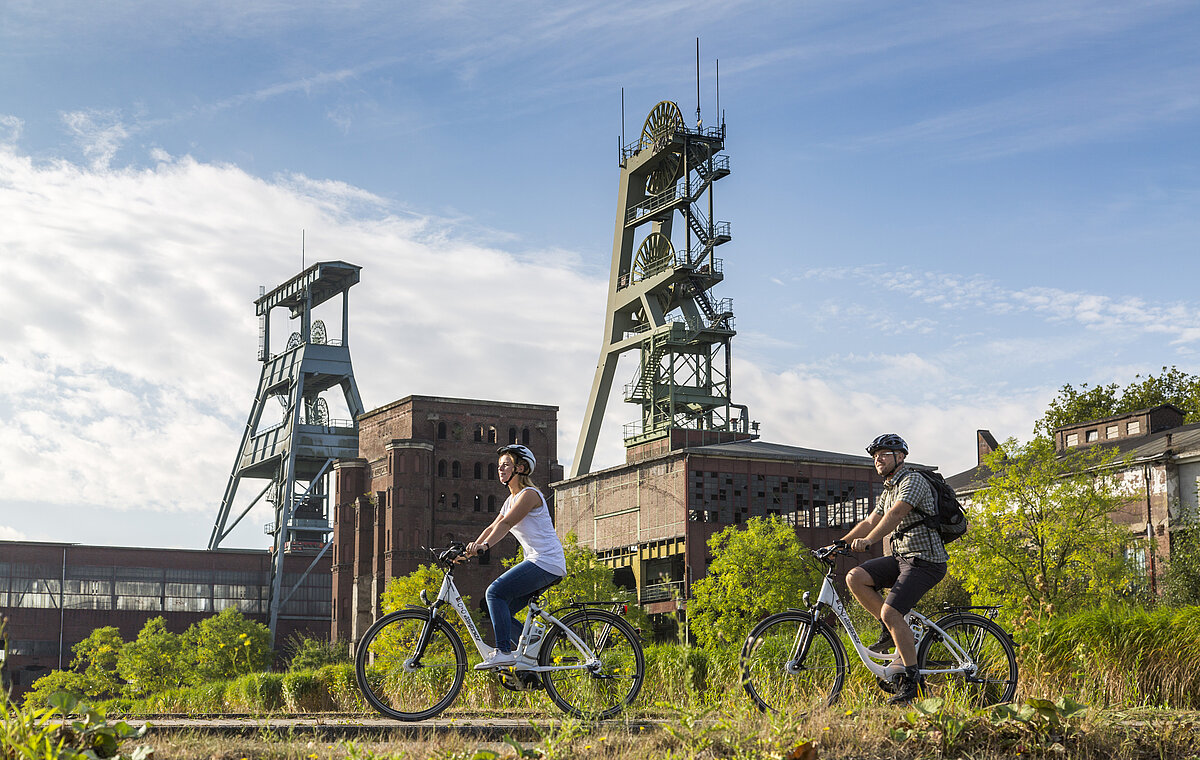 Zwei Personen auf Pedelecs fahren hintereinander über das Gelände der Zeche Ewald. Im Hintergrund sind die drei Fördergerüste zu sehen.