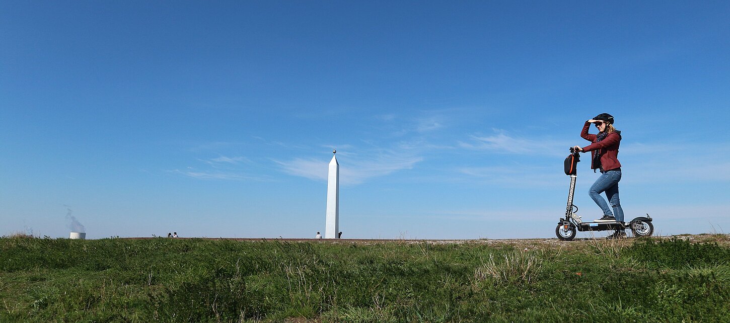 Eine junge Frau steht auf einem E-Scooter auf dem Plateau der Sonnenuhr, deren Obelisk im Hintergrund zu sehen ist und blickt in die Ferne.