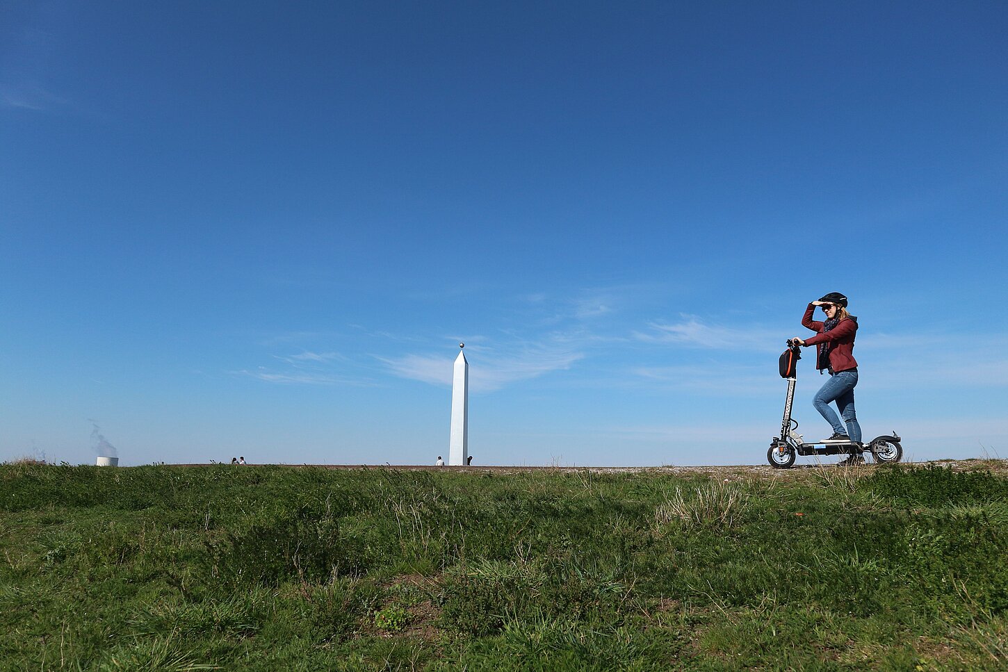 Eine junge Frau steht auf einem E-Scooter auf dem Plateau der Sonnenuhr, deren Obelisk im Hintergrund zu sehen ist und blickt in die Ferne.