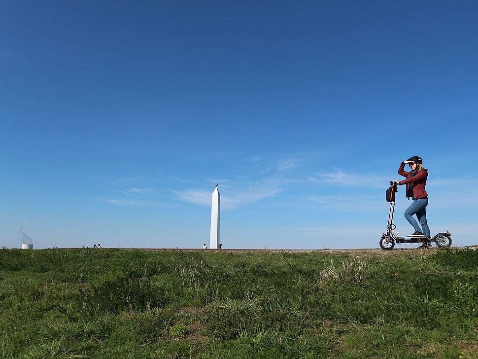 Eine junge Frau steht auf einem E-Scooter auf dem Plateau der Sonnenuhr, deren Obelisk im Hintergrund zu sehen ist und blickt in die Ferne.