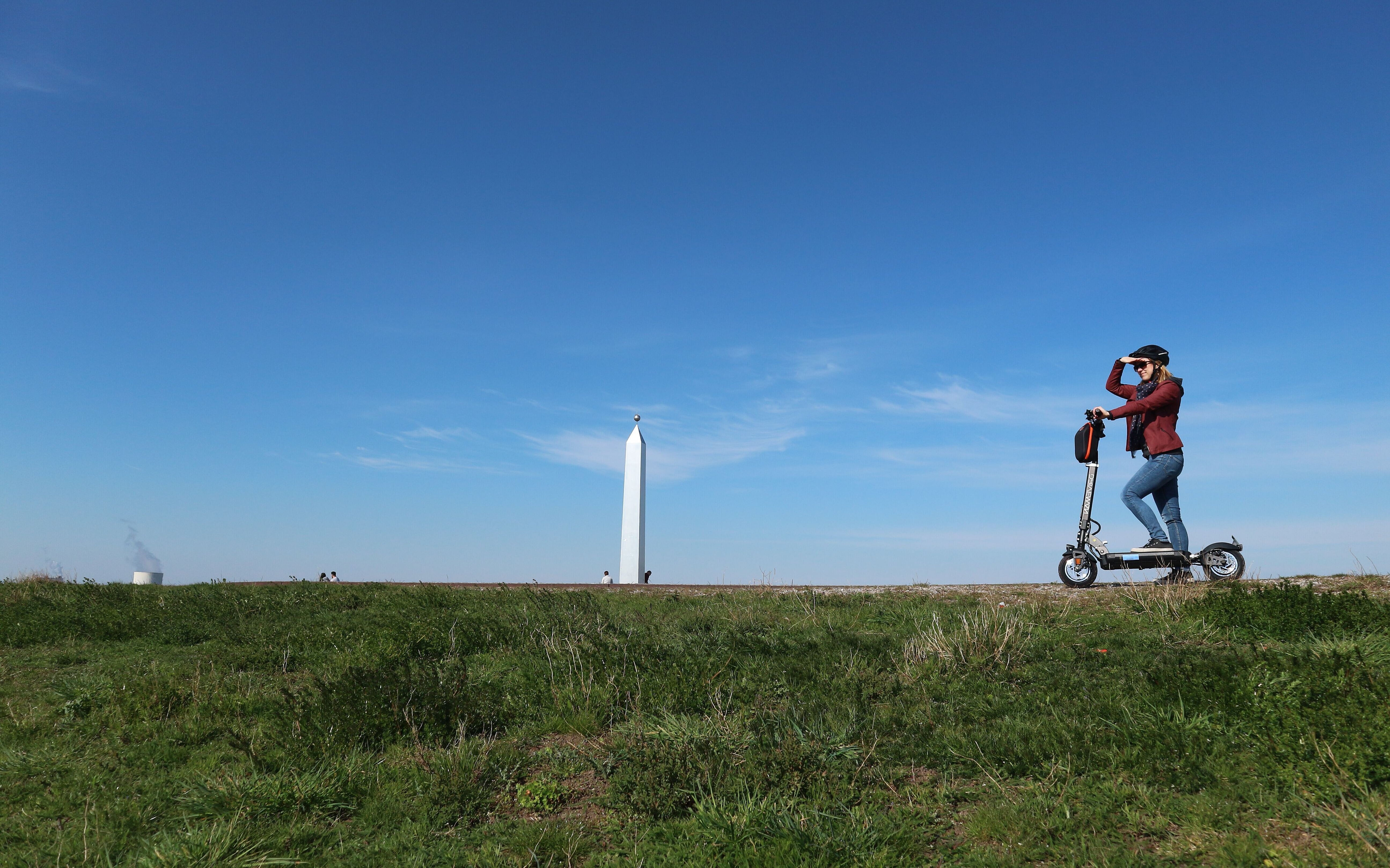 Eine junge Frau steht auf einem E-Scooter auf dem Plateau der Sonnenuhr, deren Obelisk im Hintergrund zu sehen ist und blickt in die Ferne.