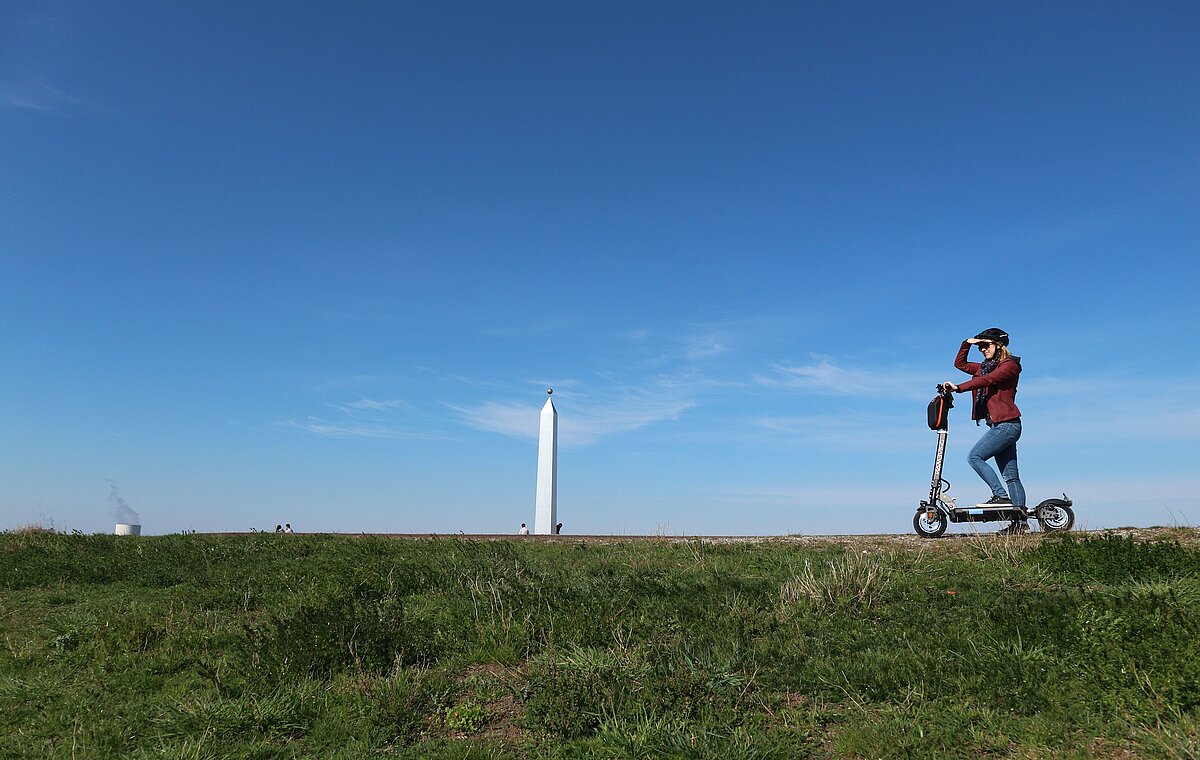 Eine junge Frau steht auf einem E-Scooter auf dem Plateau der Sonnenuhr, deren Obelisk im Hintergrund zu sehen ist und blickt in die Ferne.