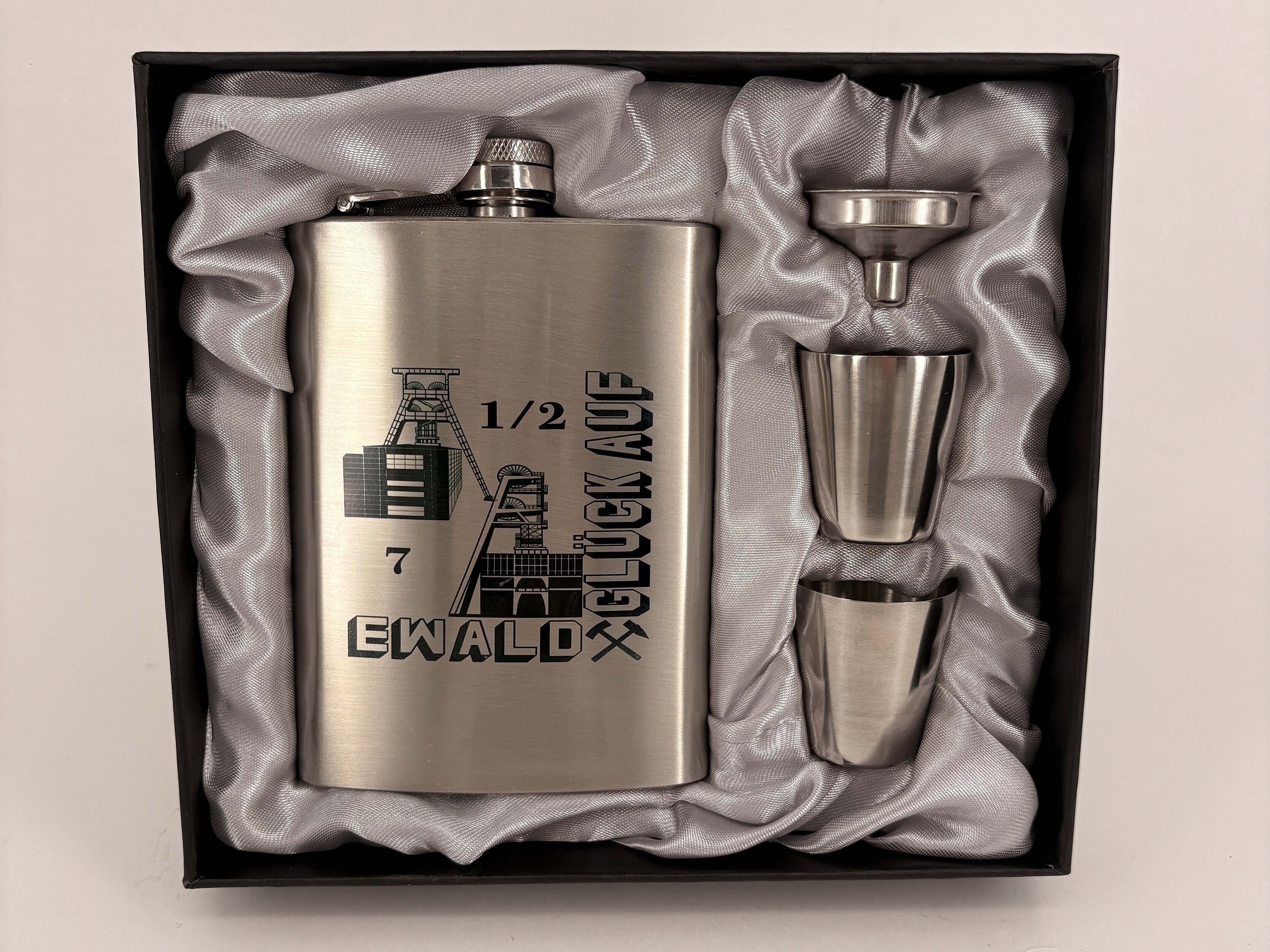 Auf dem Bild liegt ein silberner Flachmann aus Metall sowie zwei Schnapspinnchen in einer Kiste.