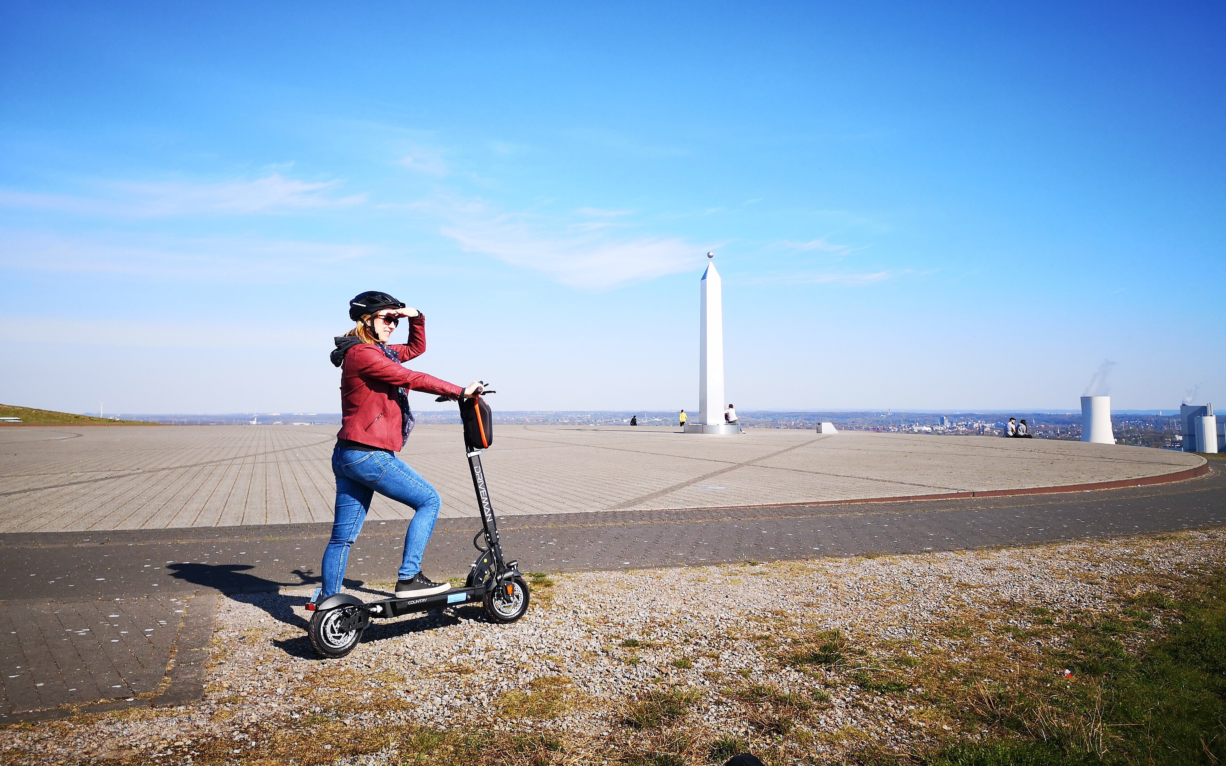 Das Bild zeigt eine Frau auf einem E-Scooter welche in die Ferne blickt, im Hintergrund ist die Sonnenuhr auf der Halde Hoheward zu sehen