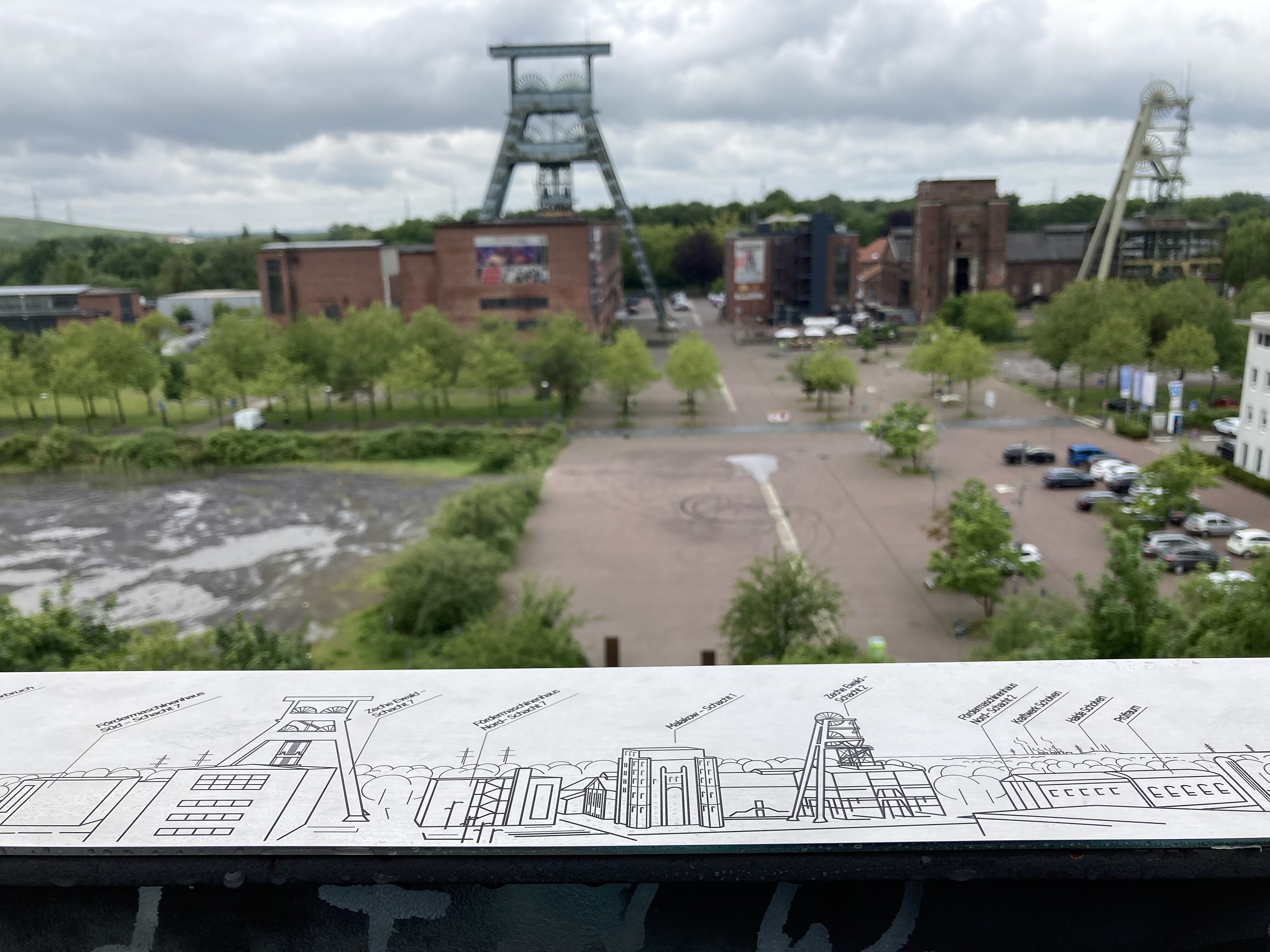 Das Foto ist von einer Aussichtplattform mit Blicke auf die Zeche Ewald fotografiert. Im Vordergrund ist eine Metallplatte zu sehen, auf welche zeigt, was von der Aussichtsplattform zu sehen ist.