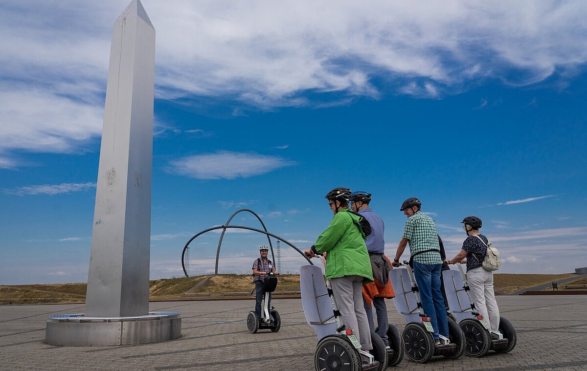 Eine Gruppe von Personen auf Segways steht neben dem Obelisken der Sonnenuhr auf der Halde Hoheward. Im Hintergrund sind die Stahlbögen des Horizontobservatorium zu sehen.