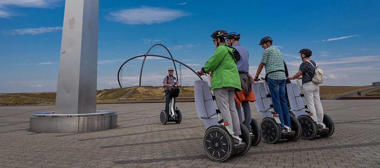 Eine Gruppe von Personen auf Segways steht neben dem Obelisken der Sonnenuhr auf der Halde Hoheward. Im Hintergrund sind die Stahlbögen des Horizontobservatorium zu sehen.