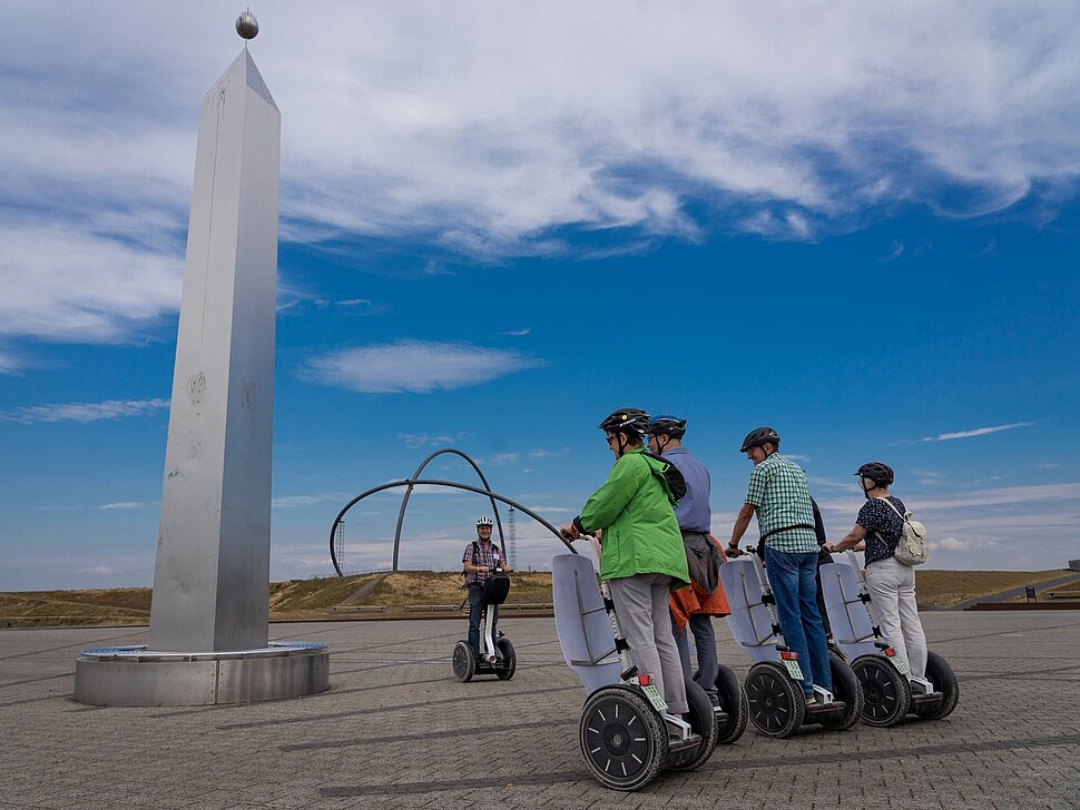 Eine Gruppe von Personen auf Segways steht neben dem Obelisken der Sonnenuhr auf der Halde Hoheward. Im Hintergrund sind die Stahlbögen des Horizontobservatorium zu sehen.