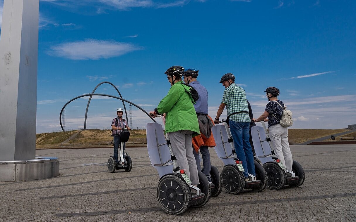Eine Gruppe von Personen auf Segways steht neben dem Obelisken der Sonnenuhr auf der Halde Hoheward. Im Hintergrund sind die Stahlbögen des Horizontobservatorium zu sehen.