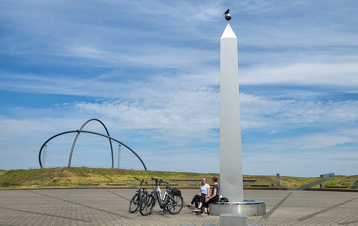 Zwei Frauen sitzen auf der Bank rund um den Obelisken der Sonnenuhr auf der Halde Hoheward. Fahrräder stehen daneben.