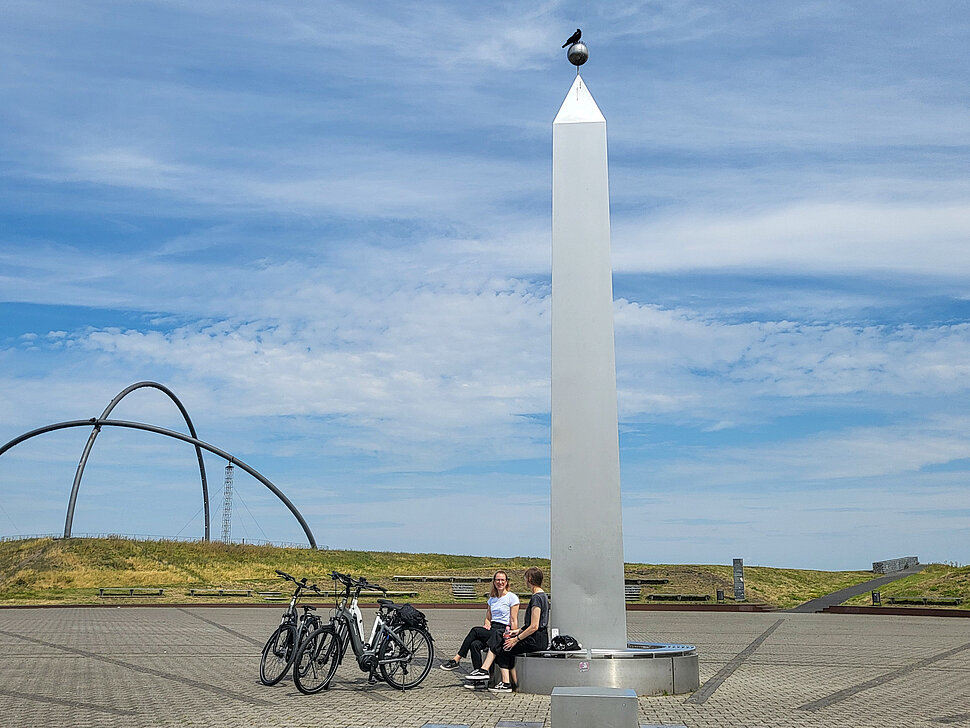 Zwei Frauen sitzen auf der Bank rund um den Obelisken der Sonnenuhr auf der Halde Hoheward. Fahrräder stehen daneben.