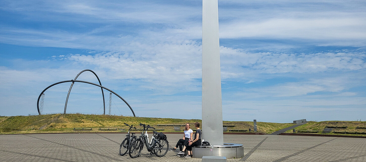 Zwei Frauen sitzen auf der Bank rund um den Obelisken der Sonnenuhr auf der Halde Hoheward. Fahrräder stehen daneben.