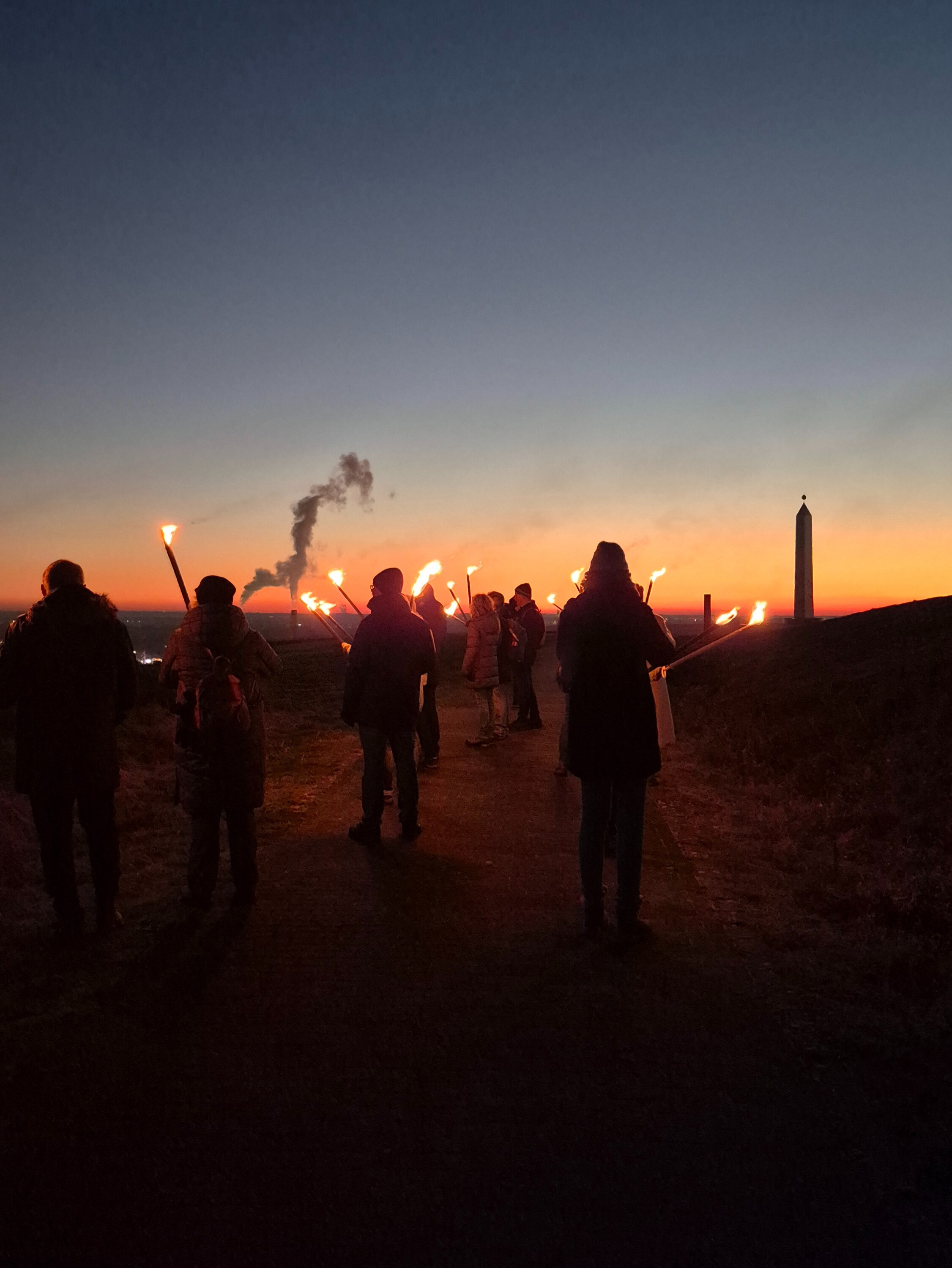 Eine Gruppe Menschen mit Fackeln läuft im Sonnenuntergang Richtung Sonnenuhr der Halde Hoheward.